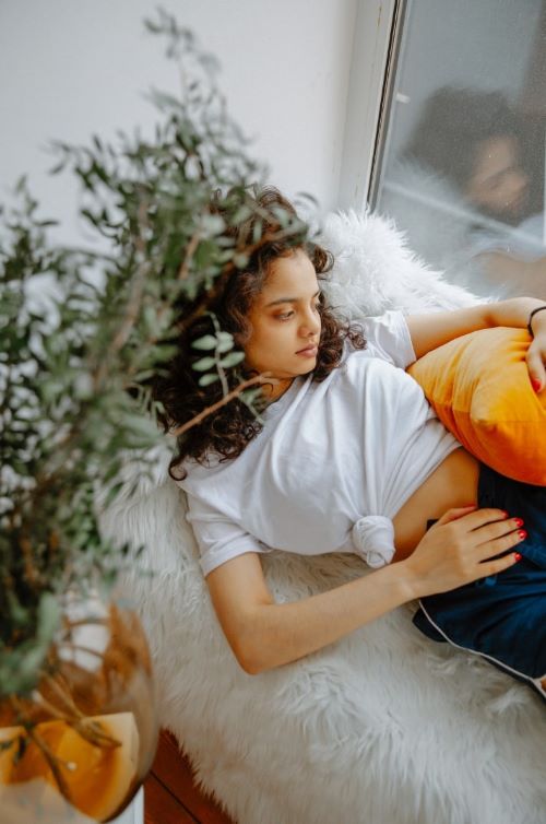 young woman lying next to window gazing out holding orange cushion over hip with one hand